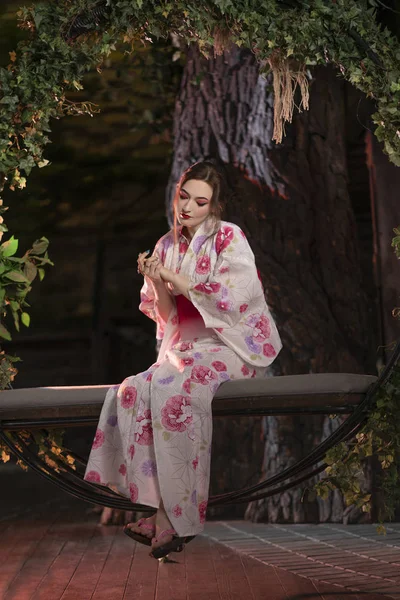 a beautiful light-skinned girl with Japanese make-up and in Japanese clothes sits on a swing, twisted with green leaves, alone at night