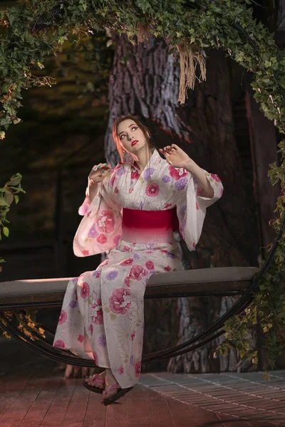a beautiful light-skinned girl with Japanese make-up and in Japanese clothes sits on a swing, twisted with green leaves, alone at night