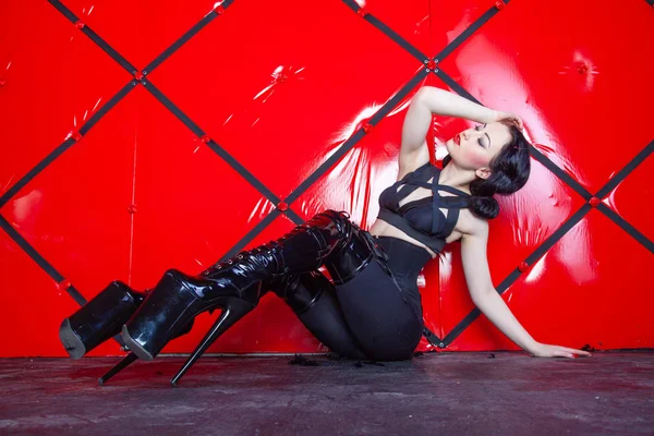 hot pin up kinky woman wearing black harness and shiny fetish thigh high boots on red background alone