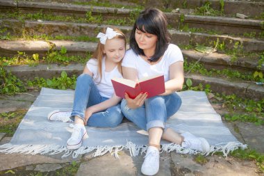 şirin anne ve tatlı kızı bir battaniye var otur ve ormanın eski çimenli adımları önünde kırmızı bir kitapta okumuştum