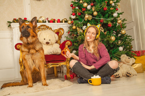 beautiful young girl in a warm sweater and yellow headphones with cat ears sitting with a Cup of cocoa with marshmallow next to a shepherd dog on the background of the Christmas tree on the floor