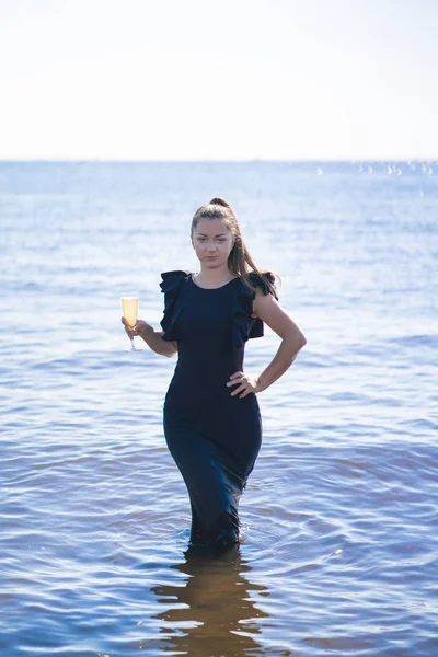 黒い長いイブニング ドレス シャンパン立って水のガラスでポーズ美しい少女 — ストック写真