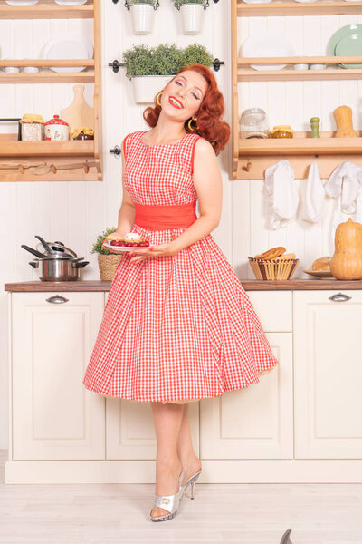 beautiful red-haired pinup smiling happily girl posing in a retro red dress in the kitchen alone