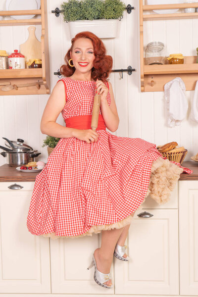 beautiful red-haired pinup smiling happily girl posing in a retro red dress in the kitchen alone