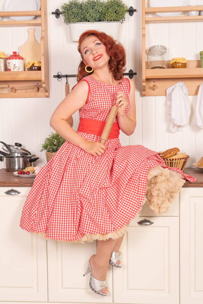 beautiful red-haired pinup smiling happily girl posing in a retro red dress in the kitchen alone