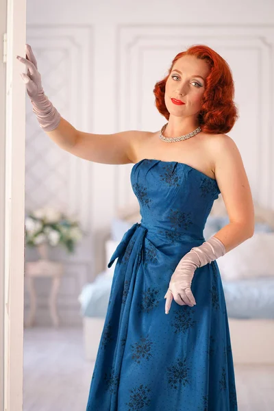 a luxurious pin up lady dressed in a blue vintage dress stands in the doorway of her bedroom and invites you to come inside