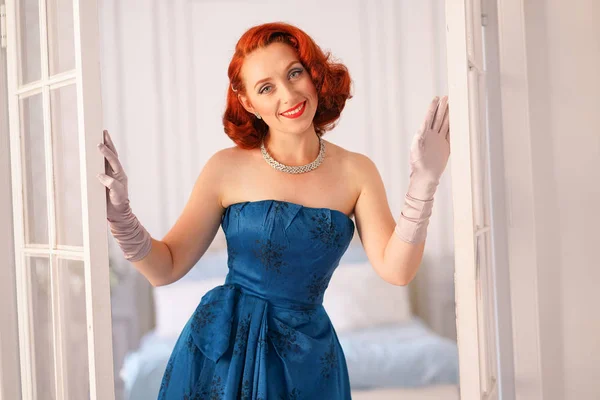 a luxurious pin up lady dressed in a blue vintage dress stands in the doorway of her bedroom and invites you to come inside