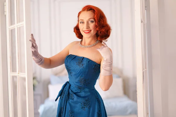 a luxurious pin up lady dressed in a blue vintage dress stands in the doorway of her bedroom and invites you to come inside