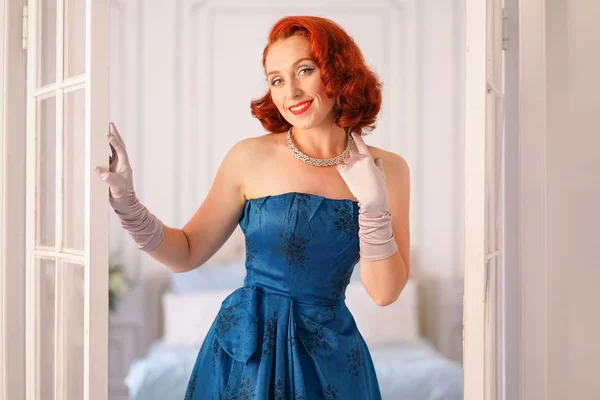 a luxurious pin up lady dressed in a blue vintage dress stands in the doorway of her bedroom and invites you to come inside