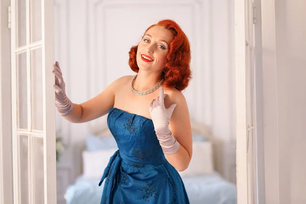 a luxurious pin up lady dressed in a blue vintage dress stands in the doorway of her bedroom and invites you to come inside