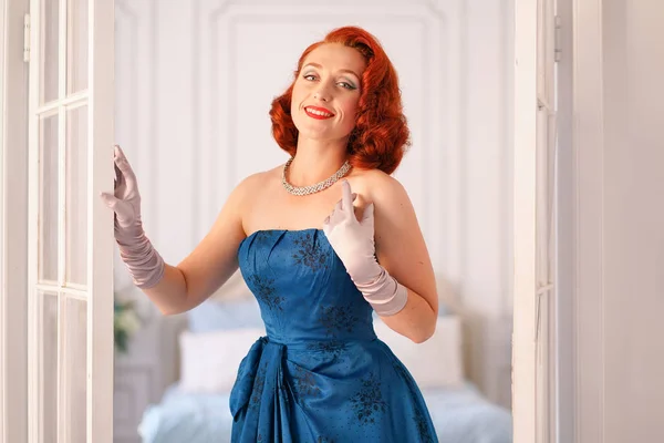 a luxurious pin up lady dressed in a blue vintage dress stands in the doorway of her bedroom and invites you to come inside