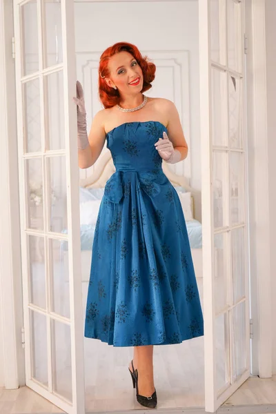 a luxurious pin up lady dressed in a blue vintage dress stands in the doorway of her bedroom and invites you to come inside