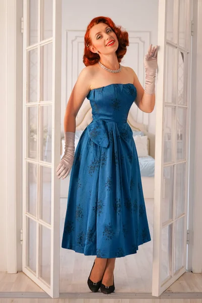 a luxurious pin up lady dressed in a blue vintage dress stands in the doorway of her bedroom and invites you to come inside