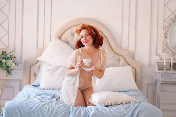 豪華なピン女性を彼女の寝室でポーズ ベージュ年代物の下着に身を包んだし 朝食の紅茶のカップを持っています — ストック写真