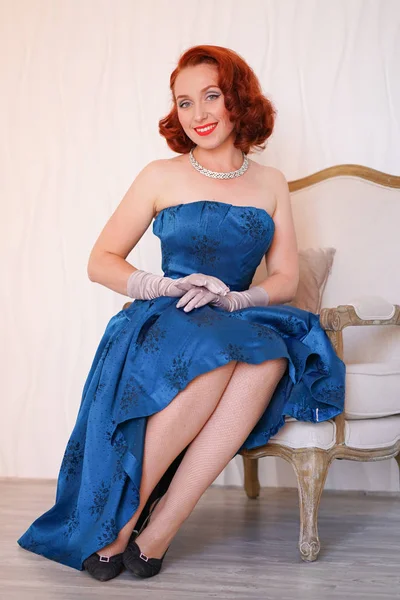 mannered beautiful vintage woman dressed in blue retro dress sitting on a chair alone and smiling on white background