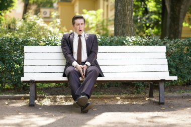 yorgun bir adam bir takım elbise içinde ofis sol ve parka gitti. o yalnız bir beyaz bankta oturan ve esnemek.
