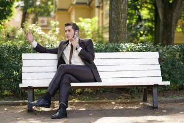 iş takım elbiseli bir genç adam ofis sol ve parka geldi. o yalnız bir beyaz bankta oturur ve telefonda konuşuyor