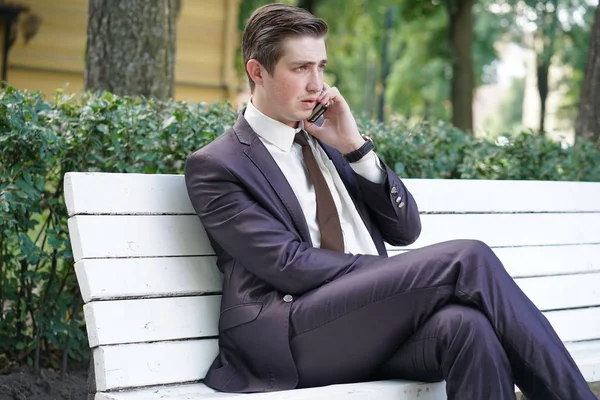 a young man in a business suit left the office and came to the Park. he sits on a white bench alone and talks on the phone