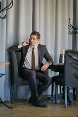 Genç bir adam bir siyah takım elbise, beyaz gömlek ve kravat sakince bir kafede bir masada oturan bir cep telefonuyla konuşuyor