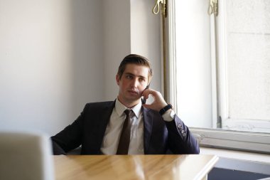 Genç bir adam bir siyah takım elbise, beyaz gömlek ve kravat sakince bir kafede bir masada oturan bir cep telefonuyla konuşuyor