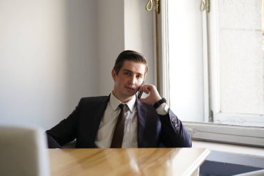 Genç bir adam bir siyah takım elbise, beyaz gömlek ve kravat sakince bir kafede bir masada oturan bir cep telefonuyla konuşuyor