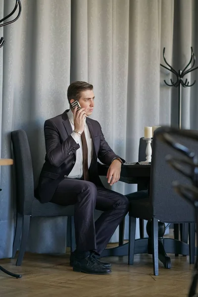 a young man in a black business suit, white shirt and tie calmly talking on a cell phone, sitting in a cafe at a table