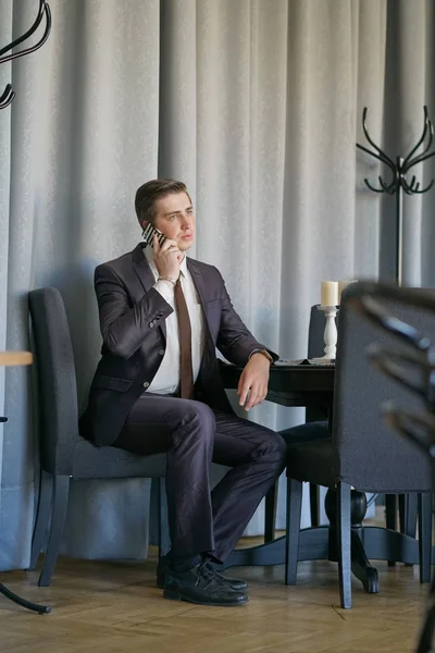 一个穿着黑色西装 白衬衫和领带的年轻人 坐在一张桌子上的咖啡馆里 冷静地用手机说话 — 图库照片