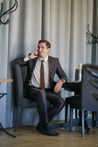 a young man in a black business suit, white shirt and tie calmly talking on a cell phone, sitting in a cafe at a table