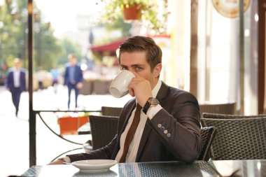 Genç bir adam bir siyah takım elbise, beyaz gömlek ve kravat bir masada bir şehir sokak kafede oturan ve bir fincan kahve keyfi