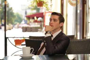 Genç bir adam bir siyah takım elbise, beyaz gömlek ve kravat bir masada bir şehir sokak kafede oturan ve bir fincan kahve keyfi