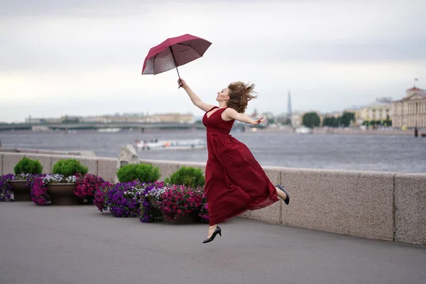 桜のロングドレスで明るく笑っている女の子がジャンプし 雨の前に薄暗い曇り空の背景に傘と付き合って — ストック写真