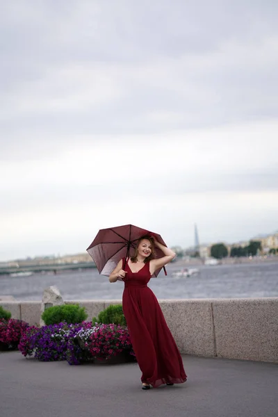 桜のロングドレスで明るく笑っている女の子がジャンプし 雨の前に薄暗い曇り空の背景に傘と付き合って — ストック写真
