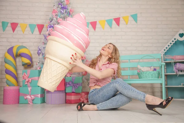 cute laughing slim girl in shirt and jeans posing with her favorite huge pink ice cream in a waffle Cup and enjoying life on the background of white Christmas tree with gifts