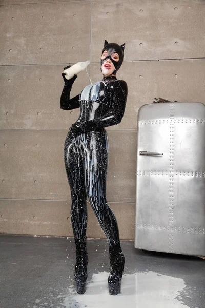 hungry cat woman with mask helmet with a bottle of milk pours it over a black latex catsuit on a silver fridge background in her grey loft apartment