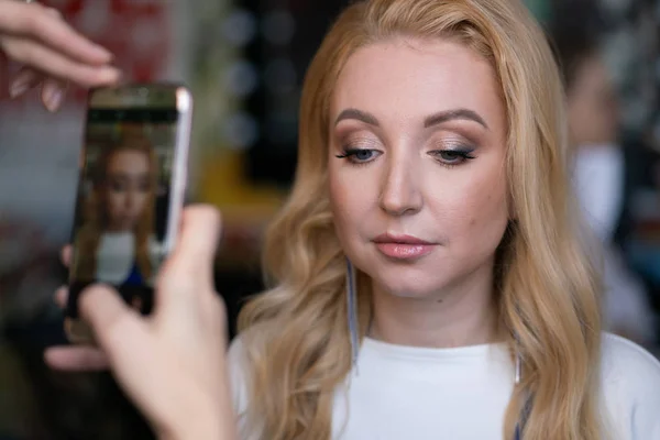 woman holding a phone in arms and taking pictures of a pretty adult friend with holiday professional make-up and hair style