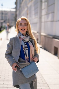 bej renkli kareli atkı gülümsüyor ve şehrin çevresinde yürüyüşler ile trendy gri paltolu uzun saçlı Sevimli sarışın kız bir turist gibi ve güneşli bir bahar günü sahiptir