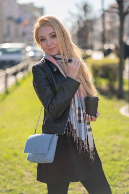 Sevimli sarışın kadın ceket damalı bir bej eşarp ile onun elinde bir fincan kahve ile yeşil şehir etrafta dolaşıyor