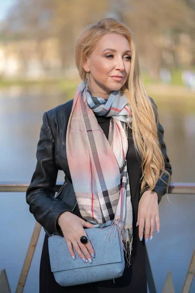 lovely blonde woman in a leather jacket, black dress and with a checkered beige scarf walks around the City and enjoys life on the positive