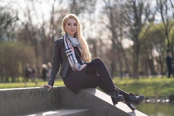 lovely blonde woman in a leather jacket, black dress and with a checkered beige scarf walks around the City and enjoys life on the positive