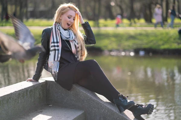 lovely blonde woman in a leather jacket, black dress and with a checkered beige scarf walks around the City and enjoys life on the positive