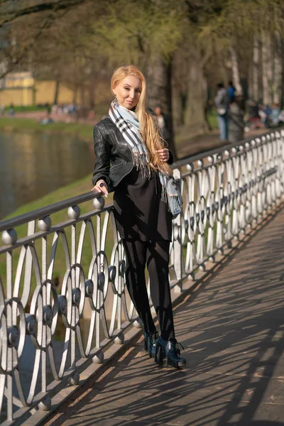 lovely blonde woman in a leather jacket, black dress and with a checkered beige scarf walks around the City and enjoys life on the positive