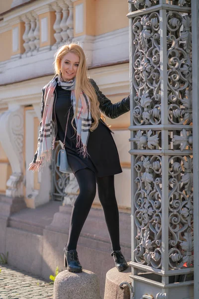 lovely blonde woman in a leather jacket, black dress and with a checkered beige scarf walks around the City and enjoys life on the positive