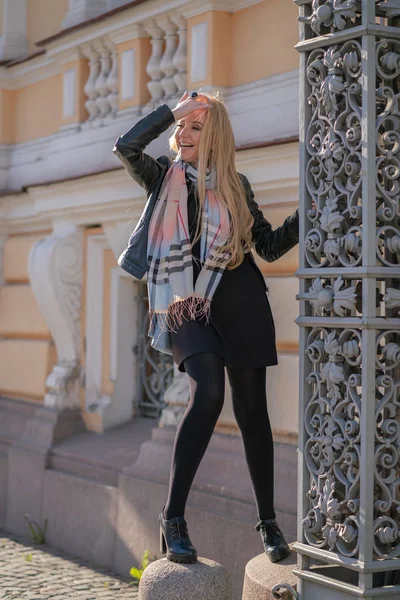 lovely blonde woman in a leather jacket, black dress and with a checkered beige scarf walks around the City and enjoys life on the positive