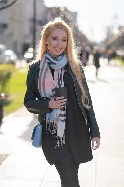 cute blonde woman in a jacket with a checkered beige scarf walks around the green city with a Cup of coffee in her hands