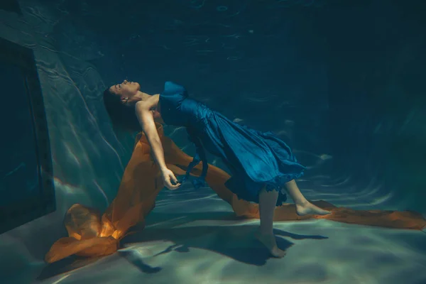 cute sporty female swims underwater as a free diver in a blue evening dress alone