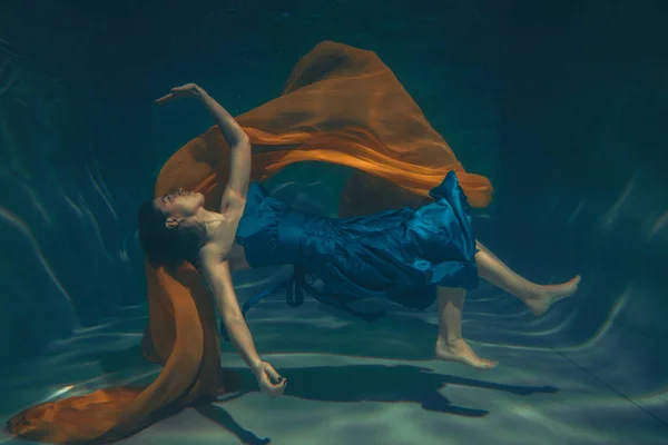 cute sporty female swims underwater as a free diver in a blue evening dress alone