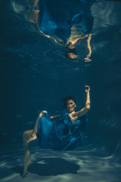 cute sporty female swims underwater as a free diver in a blue evening dress alone