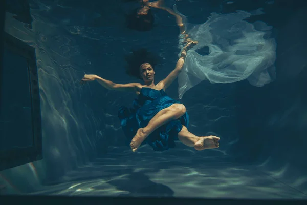 cute sporty female swims underwater as a free diver in a blue evening dress alone