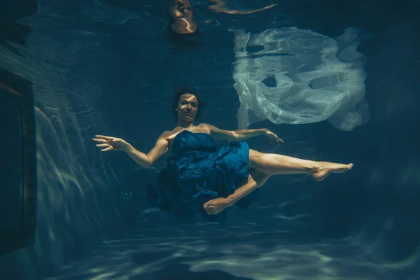 cute sporty female swims underwater as a free diver in a blue evening dress alone