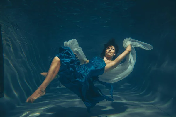 cute sporty female swims underwater as a free diver in a blue evening dress alone